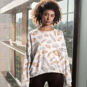 Puede incluir: Una sudadera blanca con un patrón de diferentes panes y pasteles. La modelo lleva leggings negros y zapatillas blancas.