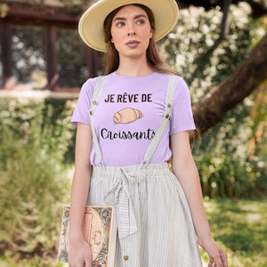 Op de afbeelding: Een vrouw draagt een lichtpaarse t-shirt met de tekst "Je rêve de croissants" en een tekening van een croissant. Ze draagt ook een witte hoed en een lichtbruin-wit gestreepte rok.