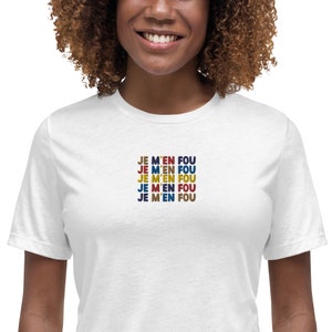 Puede incluir: Camiseta blanca con la frase "JE M'EN FOU" repetida cinco veces en un diseño apilado. El texto está en una fuente retro con diferentes colores para cada repetición.
