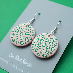 May include: Pink and green leopard print wooden earrings. The earrings are round and have a silver hook.