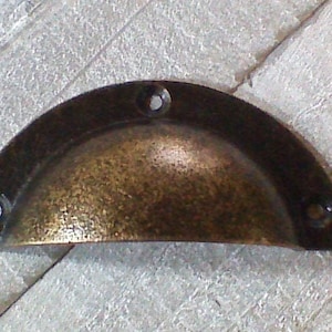 Op de afbeelding: Een halfronde, antieke messing kastgreep met een gehamerde afwerking en vier schroefgaten.