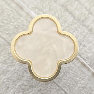 May include: A gold-coloured, four-leaf clover-shaped decorative item with a cream-coloured, marbled centre. The design has a smooth, polished finish. Likely a decorative element for furniture or jewellery.