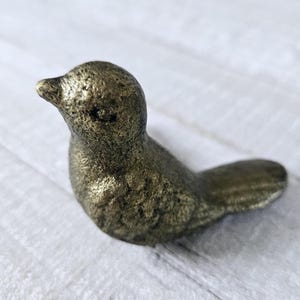 May include: A small, gold-colored cast iron bird figurine. The bird is facing to the right and has a long tail.
