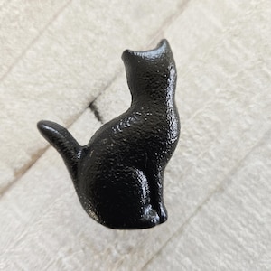 Könnte beinhalten: Eine schwarze Katzenfigur mit strukturierter Oberfläche, die aufrecht sitzt und ihren Schwanz gekrümmt hat. Die Katze ist von hinten zu sehen und zeigt ihren Rücken und die Form ihrer Ohren. Die Figur steht auf einer weißen, strukturierten Oberfläche.