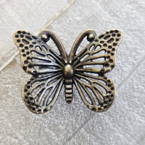 May include: A bronze-colored butterfly-shaped decorative element. The butterfly has detailed openwork wings and a textured body. The wings have a combination of solid and perforated sections. The butterfly is a decorative item.