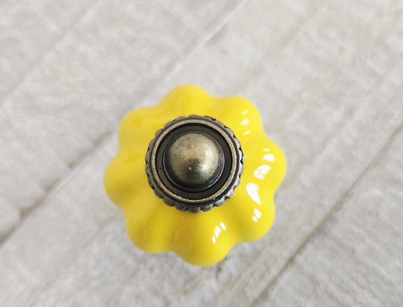 May include: A yellow ceramic cabinet knob with a scalloped edge design. The knob has a dark, antique-style metal centre. The hardware is likely for a drawer or cabinet. The knob is a decorative accent.