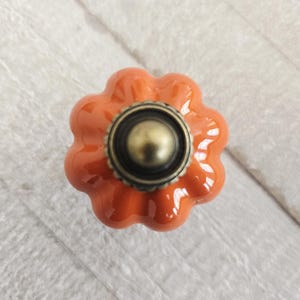 Peut inclure: Gros plan sur un bouton de meuble en céramique orange en forme de citrouille avec une base et un centre de couleur bronze. Le bouton a une finition brillante et est posé sur une surface en bois blanc.