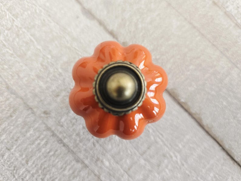 Op de afbeelding: Close-up van een oranje keramische pompoenvormige kastknop met een bronzen basis en midden. De knop heeft een glanzende afwerking en is geplaatst op een wit houten oppervlak.