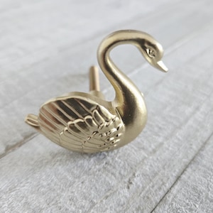 Könnte beinhalten: Ein goldfarbener schwanförmiger Schrankknauf. Der Schwan hat detaillierte Flügel- und Federdesigns. Der Knauf ist aus Metall gefertigt und zur Befestigung an einem Schrank oder einer Schublade bestimmt. Der Schwan blickt nach rechts.