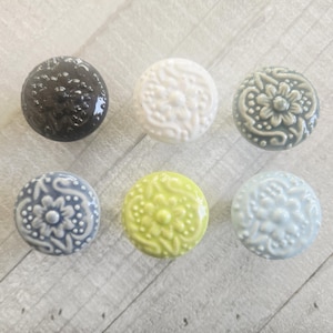 Puede incluir: Seis pomos de cerámica para armarios en varios colores: negro, blanco, gris-verdoso, azul, amarillo-verdoso y azul claro. Cada pomo presenta un diseño floral en relieve. Estos pomos decorativos son perfectos para añadir un toque de elegancia a muebles o armarios.