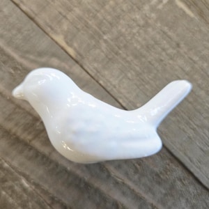 May include: A small, white ceramic bird figurine. The bird is smooth and glossy, with a rounded body and a small tail. The figurine is resting on a wooden surface.
