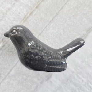 May include: A small, dark gray ceramic bird figurine. The bird has a smooth, glossy finish with textured detailing on its body. The figurine is designed in a simple, stylized form, suitable for home decor or as a decorative accent.