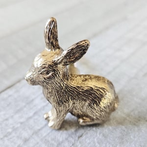 May include: A gold-colored bunny figurine with detailed fur texture. The rabbit is in a seated position with its ears upright. The figurine is likely made of metal and is a decorative item.