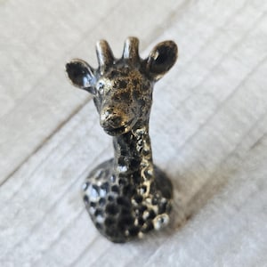 Könnte beinhalten: Bronze-farbene Giraffenfigur mit strukturierter Oberfläche. Der Kopf und Hals der Giraffe sind detailliert, mit sichtbaren Ohren und Hörnern. Die Figur ist wahrscheinlich aus Metall und hat ein rustikales, antikes Aussehen. Es ist ein dekorativer Artikel.
