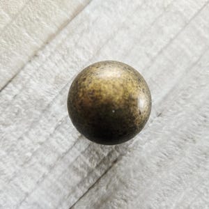 May include: A small, round, bronze-colored sphere with a speckled surface. The sphere is centered on a light gray, textured wooden surface. The sphere's color ranges from light to dark bronze.