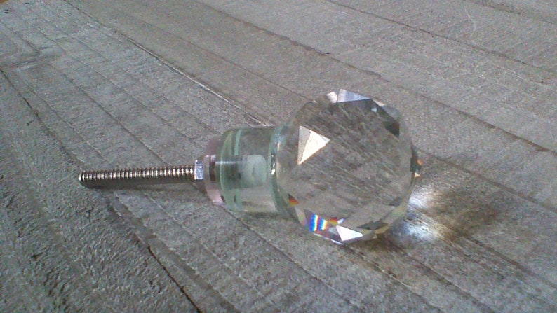 May include: A clear glass crystal cabinet knob with a silver screw. The knob has a faceted design and is shaped like a diamond.