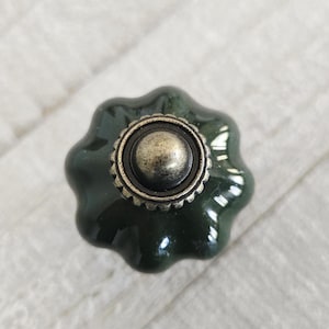 May include: A dark green ceramic cabinet knob with a flower-like shape. The knob has a metallic, antique-style centre and rim. The hardware is designed for furniture and home decor.