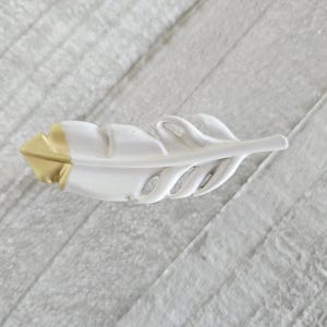 May include: A white feather-shaped decorative object with a gold-colored tip. The feather has a detailed design with carved lines and is set against a light gray, textured background.