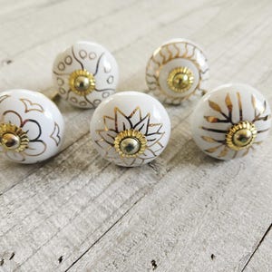 Può includere: Cinque pomelli per mobili in ceramica bianca con disegni floreali dorati e finiture dorate. I pomelli sono disposti su una superficie di legno chiaro e invecchiato. Ogni pomello presenta un motivo unico, aggiungendo un tocco decorativo.