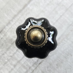 May include: A glossy black ceramic cabinet knob with a scalloped, flower-like edge and a central circular brass-toned metal accent. The hardware features a smooth, reflective finish and a raised, textured border around the center dome.