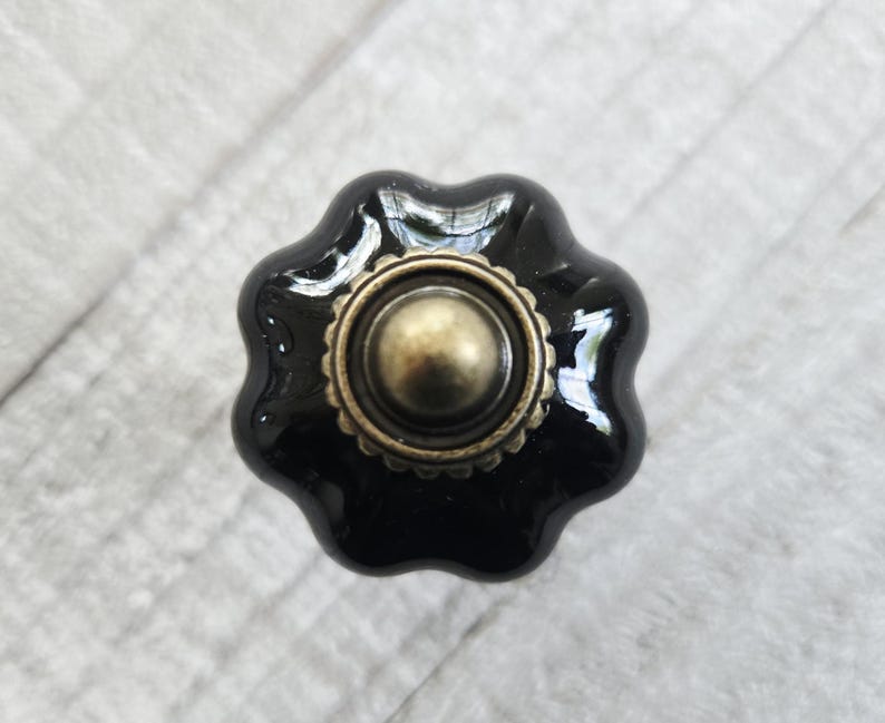 May include: A glossy black ceramic cabinet knob with a scalloped, flower-like edge and a central circular brass-toned metal accent. The hardware features a smooth, reflective finish and a raised, textured border around the center dome.
