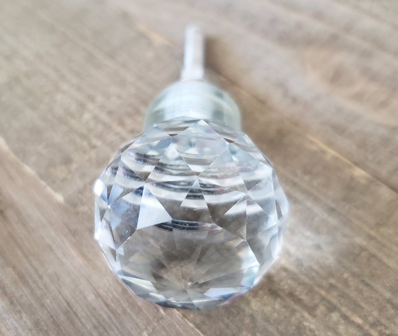 May include: A clear glass crystal knob with a faceted design. The knob has a silver metal stem.