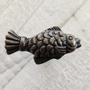 Op de afbeelding: Een bronzen visvormige kastknop met gedetailleerde schubben en vinnen. Het decoratieve hardware-stuk is ontworpen om op een vis te lijken en wordt waarschijnlijk gebruikt voor laden of kasten.