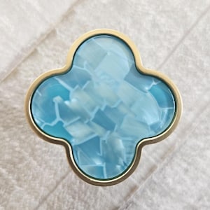 Puede incluir: Pomo de armario en forma de trébol de cuatro hojas con un borde dorado. El centro del pomo presenta un patrón azul claro, similar a un mosaico. Este herraje decorativo es adecuado para cajones y armarios.