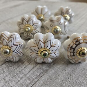 Può includere: Un set di pomelli per armadi a forma di fiore in ceramica bianca con dettagli dorati. Ogni pomello presenta un design floreale dettagliato e un centro dorato. I pomelli sono disposti su una superficie di legno chiaro.
