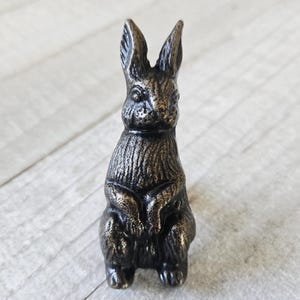 May include: A bronze-colored rabbit figurine with detailed texture. The rabbit is in a seated position with its ears upright. The figurine has a vintage or antique appearance, suitable for home decor or as a collectible item.