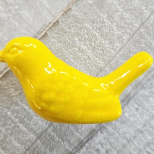 May include: A vibrant yellow ceramic bird ornament. The bird has a smooth, glossy finish and a simple, stylized design. The bird's beak and tail are clearly defined, and the overall shape is rounded and compact.
