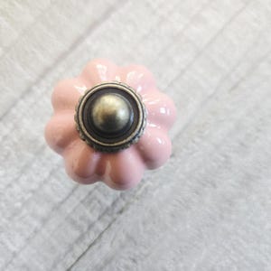 May include: A pink ceramic cabinet knob with a flower design. The knob features a bronze metal centre and a glossy finish. This decorative hardware is suitable for drawers, cabinets, and furniture.