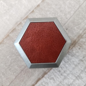 Puede incluir: Una caja hexagonal plateada con tapa de cuero rojo. La caja tiene un acabado liso y pulido.