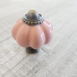 May include: A decorative cabinet knob with a pink, pumpkin-shaped ceramic body. The knob features a bronze-colored base and a matching cap with a decorative ring. The knob is designed for furniture and home decor.
