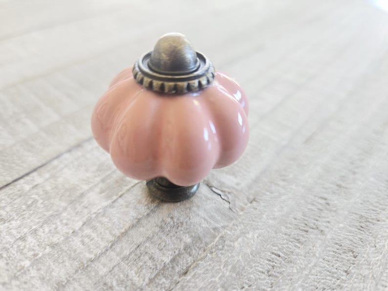 May include: A decorative cabinet knob with a pink, pumpkin-shaped ceramic body. The knob features a bronze-coloured base and a matching cap with a decorative ring. The knob is designed for furniture and home decor.