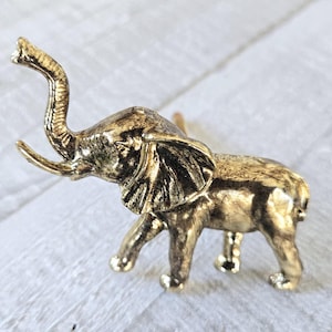 Op de afbeelding: Een gouden metalen olifantenbeeldje met zijn slurf omhoog. De olifant staat op een wit oppervlak.