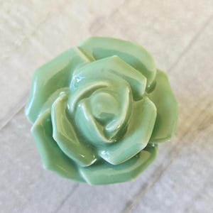 Könnte beinhalten: Eine hellgrüne Keramikrose mit glänzenden Blütenblättern steht im Mittelpunkt. Die Rose scheint ein dekorativer Gegenstand zu sein. Die Blütenblätter sind eng gewickelt und bilden ein realistisches Blumendesign. Der Hintergrund ist eine helle, strukturierte Oberfläche.