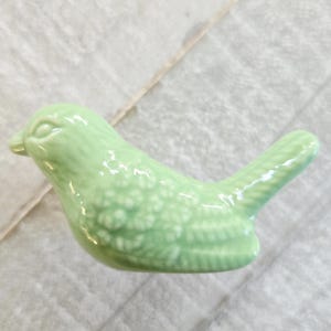Puede incluir: Figurita de pájaro de cerámica verde claro. El pájaro tiene patrones detallados de plumas en las alas y la cola. La figurita es de color sólido y tiene un acabado brillante. El pájaro es un objeto decorativo.