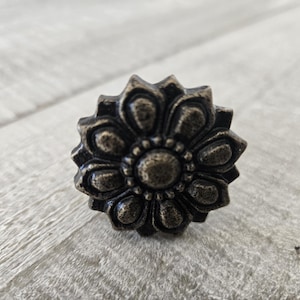 Könnte beinhalten: Ein dekorativer, blumenförmiger Knauf aus Metall mit einer antiken, abgenutzten Oberfläche. Der Knauf hat eine erhabene Mitte mit einem kreisförmigen Design und acht Blütenblättern.