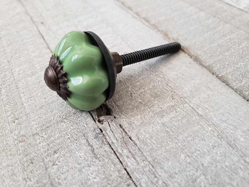 May include: A green ceramic cabinet knob with a metal base and a black screw. The knob has a rounded, pumpkin-like shape.