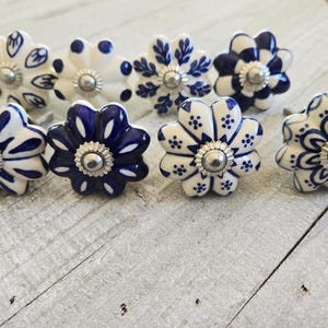 May include: A collection of eight ceramic flower-shaped cabinet knobs. The knobs feature a white base with blue floral patterns and silver-toned metal centers. The knobs are arranged on a weathered, white wooden surface.