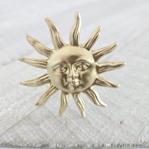 Può includere: Un oggetto decorativo dorato a forma di sole con un volto sorridente e raggi radianti. Il sole ha un'estetica vintage ed è probabilmente in metallo. L'oggetto è posto su una superficie bianca e strutturata.