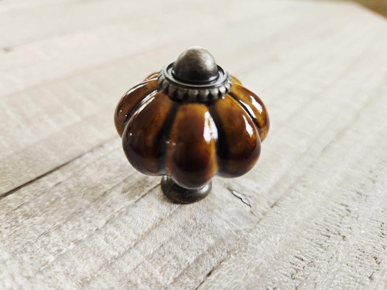 May include: A pumpkin-shaped cabinet knob in glossy brown ceramic, with a metallic silver base and top. This decorative knob is suitable for furniture. The knob's design is reminiscent of a pumpkin.