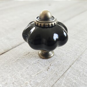 May include: A glossy black ceramic cabinet knob with a scalloped, pumpkin-like shape. The hardware features an antique brass-toned metal base and a matching decorative cap at the top, providing a contrast to the smooth, reflective dark surface.