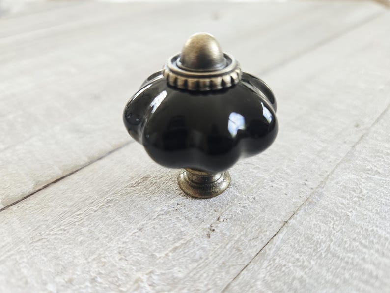 May include: A glossy black ceramic cabinet knob with a scalloped, pumpkin-like shape. The hardware features an antique brass-toned metal base and a matching decorative cap at the top, providing a contrast to the smooth, reflective dark surface.