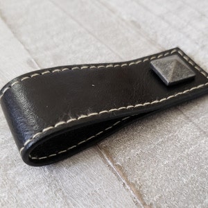 May include: A black leather drawer pull with white stitching and a square, metal accent. The pull is curved and has a stitched edge. The metal accent is a dark gray color and is square-shaped.