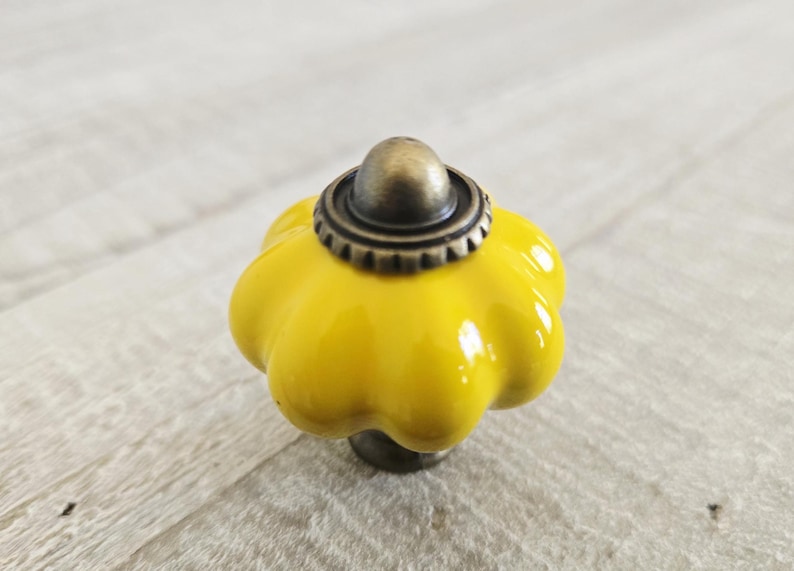May include: A bright yellow ceramic cabinet knob with a flower-like shape. The knob has a bronze-coloured base and a decorative metal cap. This decorative hardware is suitable for drawers and cabinets.