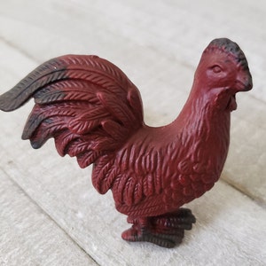 Puede incluir: Una figurita de gallo rojo con cola negra y plumas detalladas. El gallo está de pie sobre una superficie de madera blanca.
