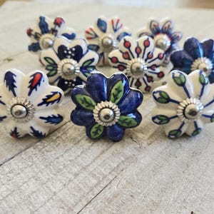 Puede incluir: Un conjunto de tiradores de armario de cerámica con forma de flor. Los tiradores presentan varios diseños florales en blanco, azul, rojo y verde. Cada tirador tiene un centro de metal plateado. Estos tiradores decorativos son ideales para muebles.