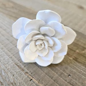 May include: A white ceramic flower with a detailed design, resembling a rose or a carnation. The flower is a decorative accent for a home or office.